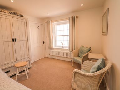 A small room with two wicker chairs a wardrobe a stool and a window with curtains at Searles in Lympstone