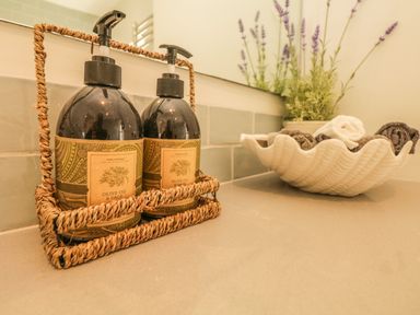 Two bottles of olive oil and sage soap in a woven holder on a countertop with rolled towels in a shell-shaped dish and lavender in a pot on the countertop at Searles in Lympstone