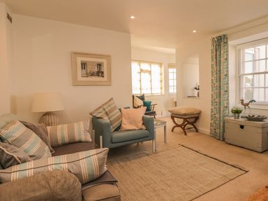 A living room with a sofa and armchair with striped pillows a lamp a framed picture on the wall a window with curtains and a chest at Searles in Lympstone