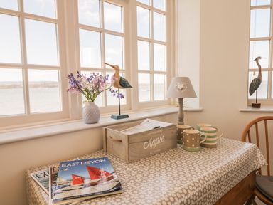 A dining table with cups, magazines, a wooden crate, a lamp, flowers, and bird figurines by large windows at Searles in Lympstone
