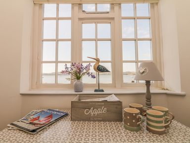 A table with mugs magazines a wooden box a lamp and a vase with flowers in front of windows showing a water view at Searles in Lympstone