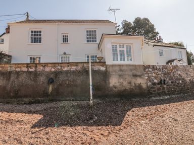 A white house with multiple windows behind a stone wall and a rocky ground in front at Searles in Lympstone