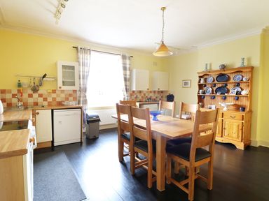 A kitchen with a wooden dining table and six chairs a wooden hutch with blue and white dishes a window with plaid curtains and yellow walls at Glebe House in Wells