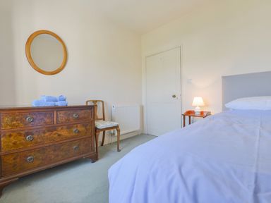 A bedroom with a bed covered with a white sheet a wooden dresser with blue towels on top a wooden chair a round wall mirror and a bedside table with a lamp at Glebe House in Wells