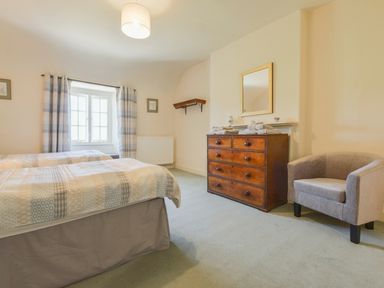 A bedroom with two single beds, a wooden dresser with folded towels, a mirror above the dresser, a gray armchair, and a window with curtains at Glebe House in Wells