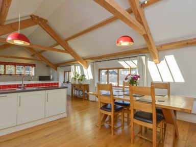 A dining area with wooden table and chairs next to large windows and an open kitchen with a sink at Old Armoury Barn Ashburton