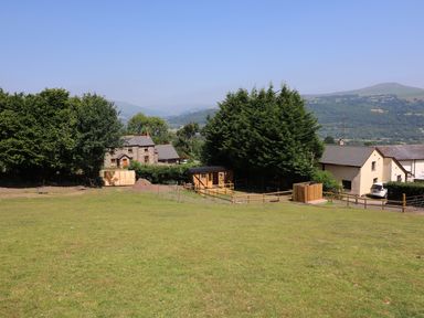 A grassy field with trees wooden sheds and stone houses with hills in the background at Railway Carriage in Govilon