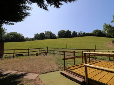 A fenced grassy field with a wooden platform and logs arranged around a fire pit at Railway Carriage in Govilon