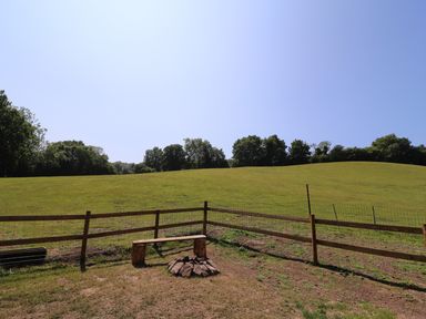 An outdoor area with a wooden bench and a fire pit enclosed by a wooden fence on a grassy hill with trees in the background at Railway Carriage in Govilon