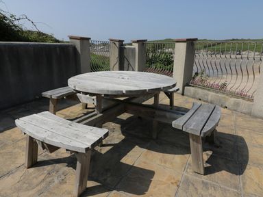 A wooden round picnic table with attached benches on a tiled patio with metal railings and a view of grassy landscape at Seashore House in Porth