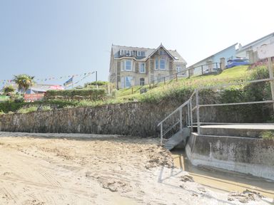 A sandy beach with steps leading up to a stone wall and a large house on a grassy hill at Seashore House in Porth