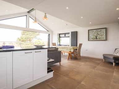 An open kitchen and dining area with a table covered by a cloth chairs and large windows at Trenant View in Wadebridge