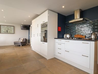 A kitchen with white cabinets and appliances near a blue tiled backsplash and a gray chair with a pillow at Trenant View in Wadebridge
