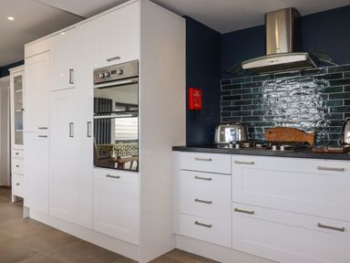 A kitchen with white cabinets a built in oven a gas stove a stainless steel hood and a fire blanket on the wall at Trenant View in Wadebridge