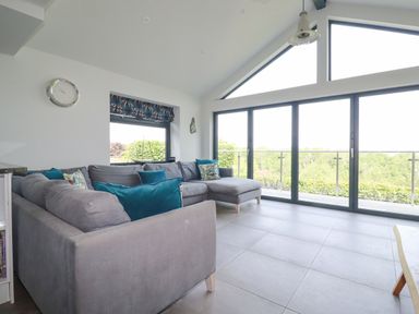 A living room with a large gray sectional sofa and glass sliding doors overlooking greenery at Trenant View in Wadebridge