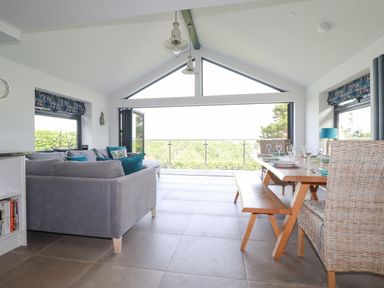 A living room with a gray sofa and a wooden dining table with chairs near large windows at Trenant View in Wadebridge