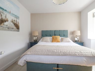 A bedroom with a bed with a tufted headboard two bedside tables with lamps and a painting on the wall at Trenant View in Wadebridge