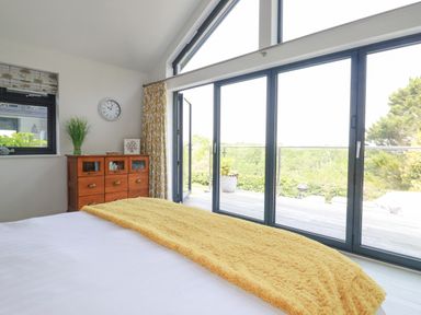 A bedroom with a bed covered with a yellow blanket a wooden dresser with decor and large glass doors opening to a balcony at Trenant View in Wadebridge