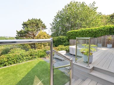 An outdoor patio area with glass and metal railings overlooking a garden with trees and shrubs at Trenant View in Wadebridge