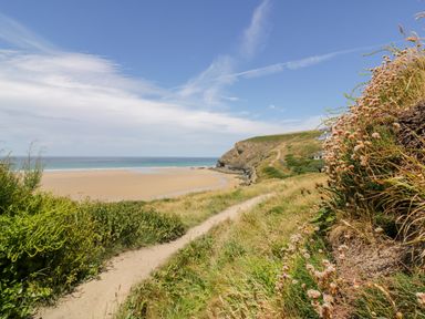 A sandy beach with a grassy path leading towards cliffs under a blue sky at Little May in Newquay