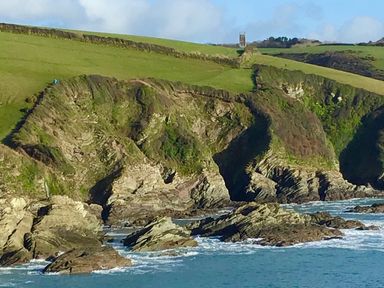 Rocky coastline with green cliffs and a tower in the distance at The Coach House at Pendower House in Fowey