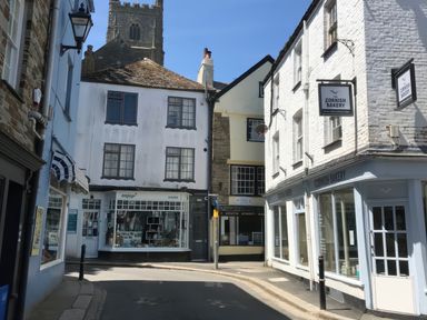 A narrow street with shops including a bakery and a gallery under a church tower in Fowey