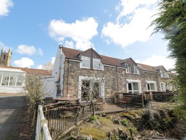 A stone cottage with multiple windows and a fenced patio area with benches and tables at Chillingham Cottage in Belford