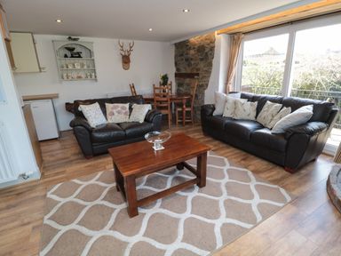 A living room with two black leather sofas with cushions a wooden coffee table a patterned rug wooden dining table and chairs and a large window at Chillingham Cottage in Belford