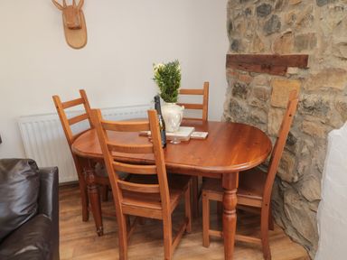 A wooden dining table with chairs a vase with flowers a bottle and a glass near a stone wall at Chillingham Cottage in Belford