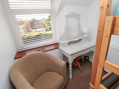 A small room with a beige armchair a white vanity table with a mirror and lamp a wooden stool and part of a wooden bunk bed at Chillingham Cottage in Belford