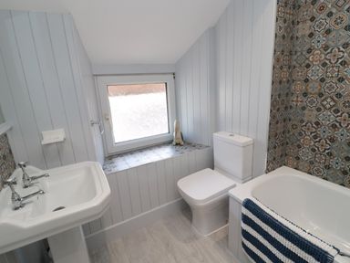A bathroom with a white sink pedestal toilet and bathtub with patterned tiles on one wall and a window with tiled sill at Chillingham Cottage in Belford