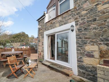 An outdoor patio area with wooden chairs and a table next to a stone building at Chillingham Cottage in Belford
