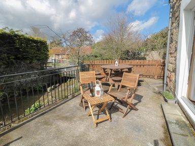 An outdoor patio with wooden chairs a small table and a round picnic table near a railing by a stream at Chillingham Cottage in Belford