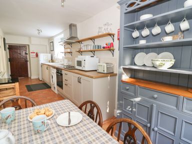 A kitchen with a blue cabinet holding plates and cups a dining table with chairs and mugs and cookies on it and a counter with a microwave toaster and sink at Pengraig near Tregaron