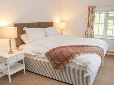 A bedroom with a bed with white bedding and a checkered throw blanket two bedside tables with lamps and a window with floral curtains at The Old Farmhouse Upper Pitts in Knighton