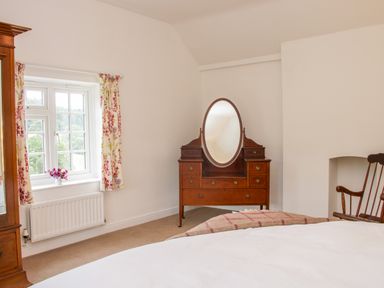A bedroom with a wooden dresser with an oval mirror a window with floral curtains a wooden rocking chair and a bed at The Old Farmhouse Upper Pitts in Knighton