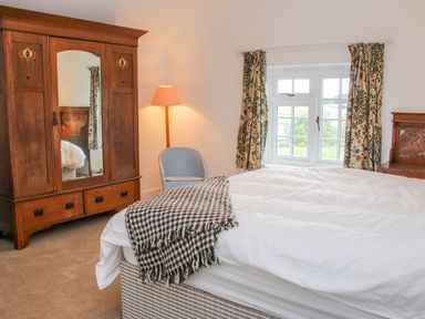 A bedroom with a bed a wooden wardrobe with mirror a blue chair floor lamp and window with patterned curtains at The Old Farmhouse Upper Pitts in Knighton