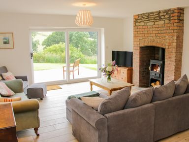 A living room with a gray sofa green armchairs a wooden coffee table flowers a brick fireplace and sliding glass doors showing a garden at The Old Farmhouse Upper Pitts in Knighton
