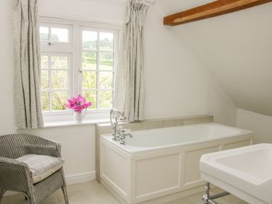 A bathroom with a bathtub near a window with curtains a wicker chair and a sink at The Old Farmhouse Upper Pitts Knighton