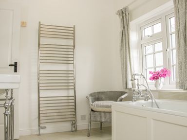 A bathroom with a towel radiator a wicker chair a bathtub and a window with curtains and flowers at The Old Farmhouse Upper Pitts in Knighton