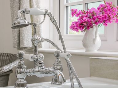 A chrome bathtub faucet with a handheld shower and pink flowers in a white vase on a windowsill at The Old Farmhouse Upper Pitts in Knighton