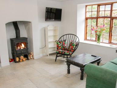 A living room with a wood stove fire a black chair with floral cushions a green sofa a wooden coffee table a white shelf and a window with flowers at The Old Farmhouse Upper Pitts in Knighton