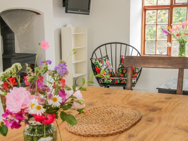 A dining area with a wooden table and flowers in a vase beside a black chair with floral cushions and a window at The Old Farmhouse Upper Pitts Knighton