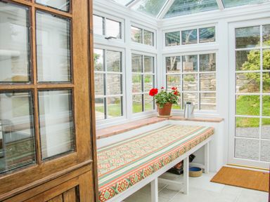 A sunroom with padded bench seating next to white framed windows and a door with a flowerpot and watering can on the windowsill at The Old Farmhouse Upper Pitts in Knighton