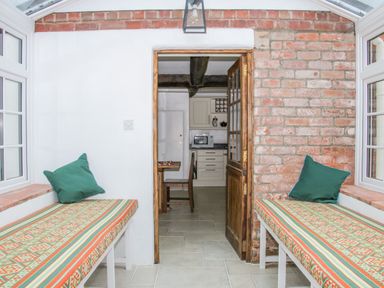 A conservatory with built in benches with cushions and a view into a kitchen through a wooden door at The Old Farmhouse Upper Pitts in Knighton