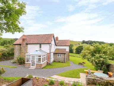 A house with a glass conservatory and garden furniture on a lawn with hills in the background at The Old Farmhouse Upper Pitts in Knighton