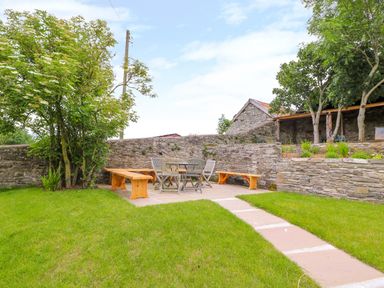 A garden with a paved seating area with wooden benches and metal chairs surrounded by stone walls and trees at The Old Farmhouse Upper Pitts in Knighton