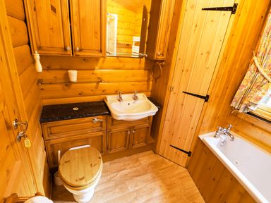 A bathroom with wooden walls and floor a toilet a sink with cabinets a bathtub and a window with floral curtains at Lodge 8 in Louth