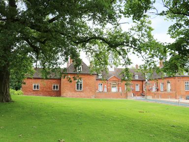 A large brick building with multiple windows and a clock tower behind a grassy lawn with tree branches overhead at Lodge 8 in Louth