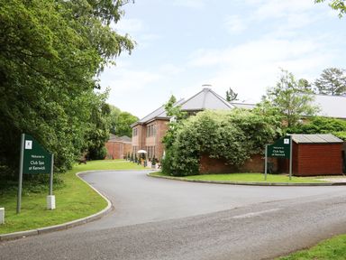Entrance road leading to a brick building surrounded by trees and greenery with welcome signs for Club Spa at Kenwick